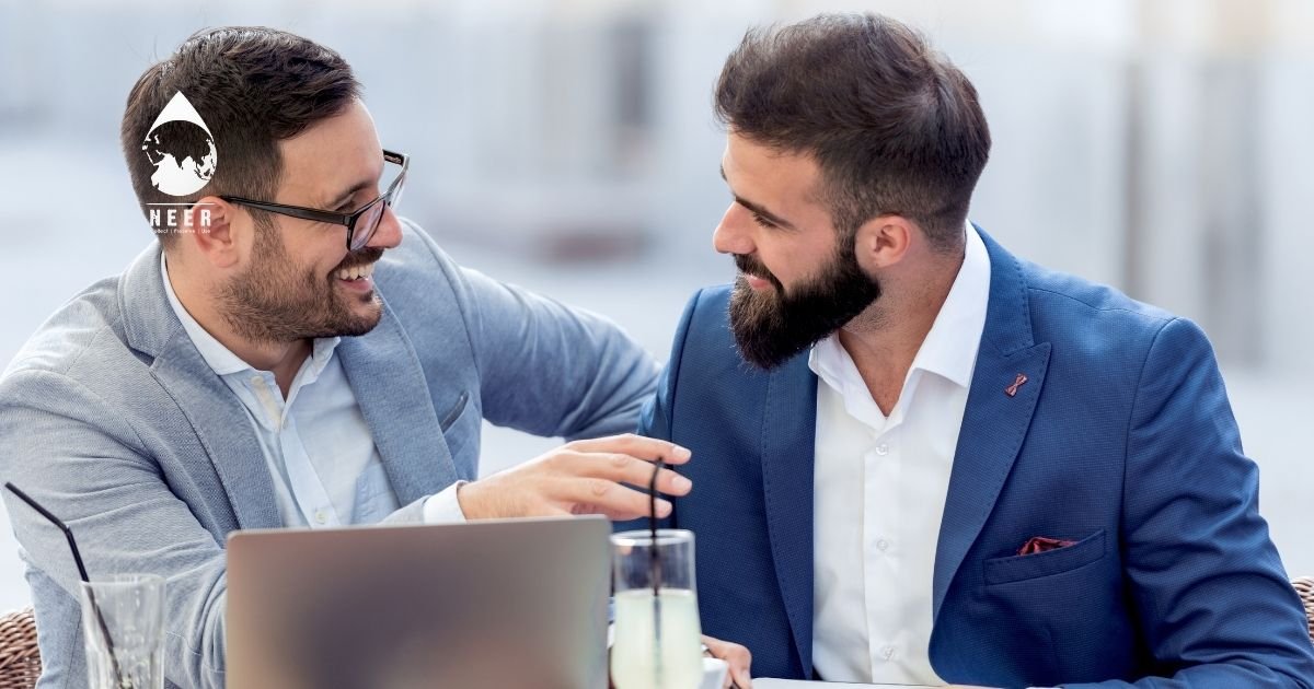 Two businessmen in light and dark blue jackets chat and smile at an outdoor caf&eacute;, laptop and drinks on the table nearby.